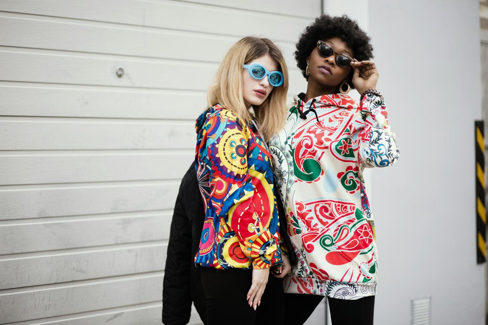 Two stylish women wearing colorful graphic hoodies and sunglasses against a modern urban backdrop.